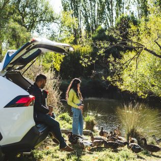 A couple enjoying a leisurely moment by a river during their Off Season road trip, standing by their car while watching ducks play. They appear relaxed, sipping coffee, and sharing a peaceful moment in the sunshine.