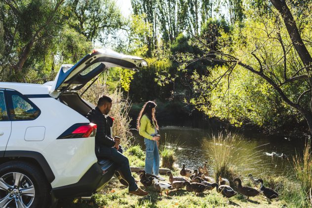 A couple enjoying a leisurely moment by a river during their Off Season road trip, standing by their car while watching ducks play. They appear relaxed, sipping coffee, and sharing a peaceful moment in the sunshine.